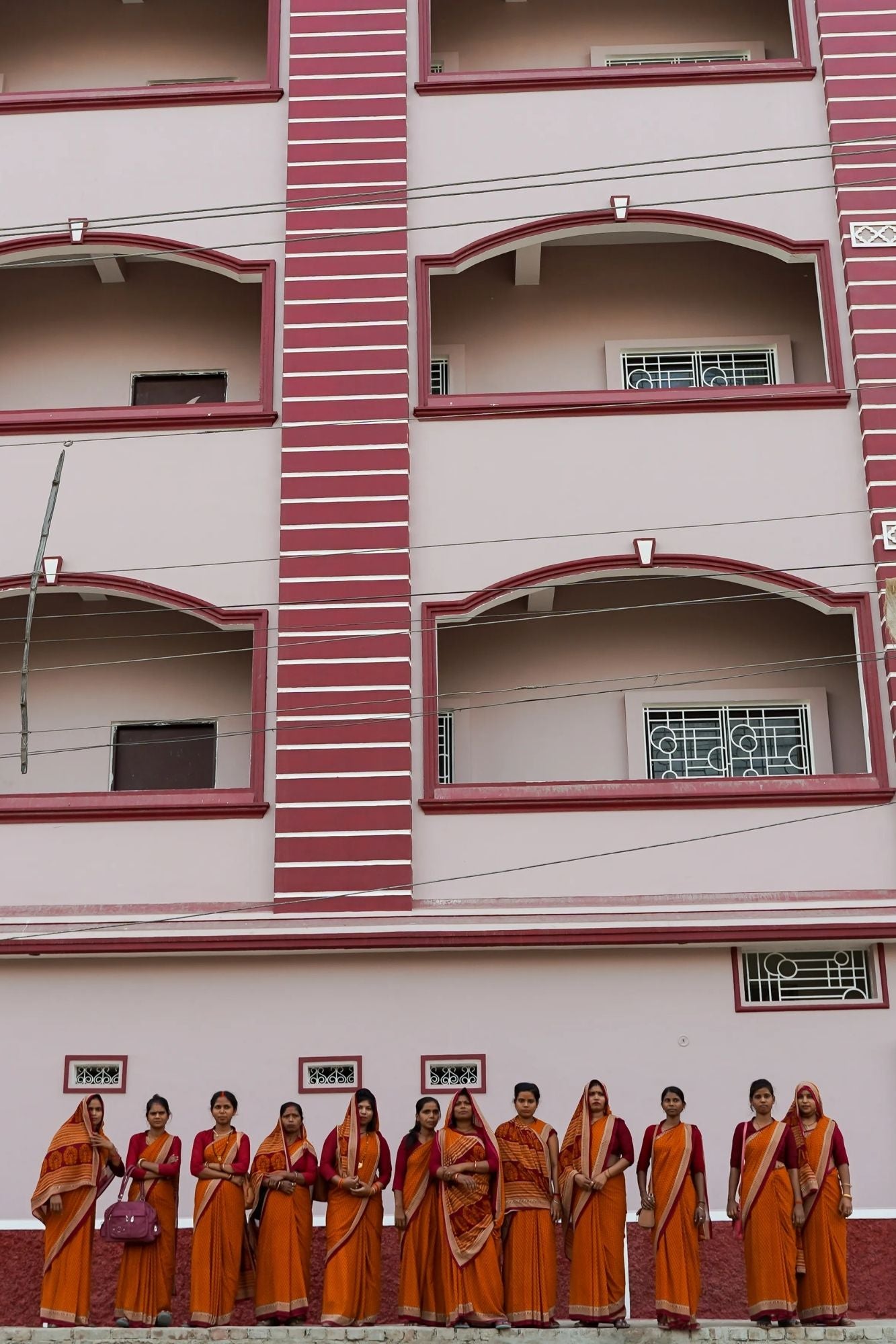 Row of women in orange sarees standing in front of a building with red and white architectural details.